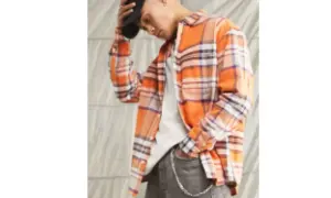 Uomo con camicia in flanella a quadri arancioni, jeans grigi e cappellino nero.