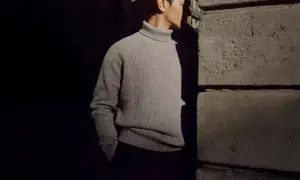 Uomo con un elegante outfit con dolcevita grigio, in piedi vicino a un muro di pietra.
