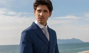 Uomo con abito blu gessato e camicia bianca, indossa una cravatta con un elegante nodo cravatta simmetrico. Sguardo pensieroso verso l'orizzonte marino.