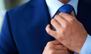 Uomo in abito blu che sistema la cravatta Windsor. Dettaglio elegante e raffinato.