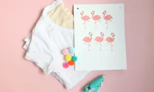 White t-shirt with colorful pom-poms, a print of pink flamingos, and a hot glue gun on a pink background.