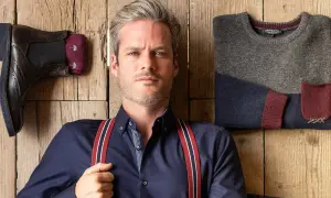 Uomo con outfit bretelle, camicia blu, stivali e maglione coordinato su fondo in legno.