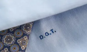 Dettaglio di una camicia azzurra con un inserto di tessuto fantasia blu e oro. Le iniziali D.G.T. sono ricamate in blu.