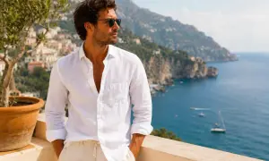 Uomo con camicia bianca in **color lino** e pantaloni beige, con vista sul mare e su un villaggio costiero.
