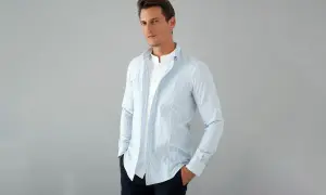 Uomo con outfit camicia a righe azzurre e bianche, indossata aperta su una t-shirt bianca. Mani nelle tasche, look casual.