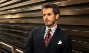 Uomo con abito blu, camicia a righe e cravatta verde con fantasia. Sguardo pensieroso.