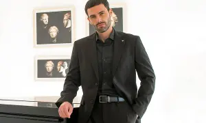 Elegante uomo con camicia invernale uomo nera sotto giacca coordinata, appoggiato a un pianoforte.