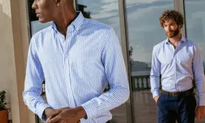 Uomo con camicia personalizzata a righe azzurre e bianche, in piedi vicino a una vetrata.