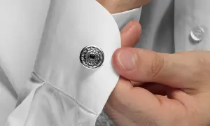 Dettaglio su come si mettono i gemelli: una mano inserisce un gemello rotondo con decorazioni argentate e una pietra nera in un polsino di camicia bianca.