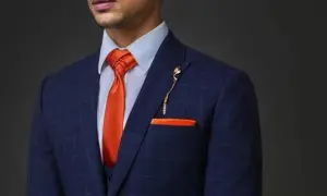 Uomo con abito blu a quadri, camicia azzurra e cravatta arancione con nodi cravatta particolari. Taschino e spilla coordinati.