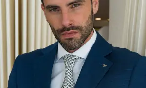 Uomo con vestito blu, camicia bianca e cravatta a fantasia. Sguardo intenso, barba curata.