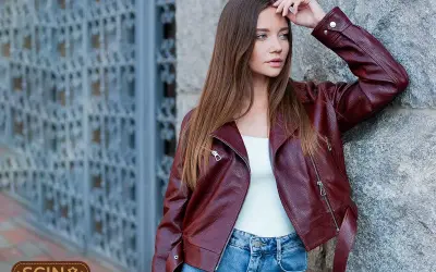 Woman in a burgundy leather jacket and blue jeans leans against a stone wall.