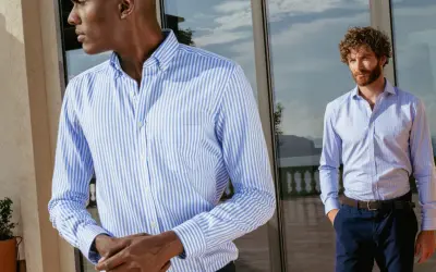 Uomo con camicia personalizzata a righe azzurre e bianche, in piedi vicino a una vetrata.