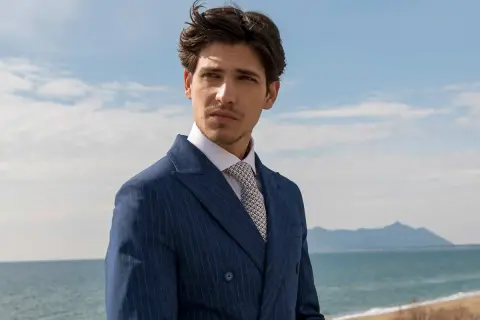 Uomo con abito blu gessato e camicia bianca, indossa una cravatta con un elegante nodo cravatta simmetrico. Sguardo pensieroso verso l'orizzonte marino.