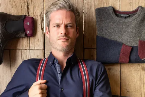 Uomo con outfit bretelle, camicia blu, stivali e maglione coordinato su fondo in legno.