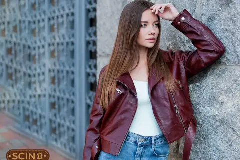 Woman in a burgundy leather jacket and blue jeans leans against a stone wall.