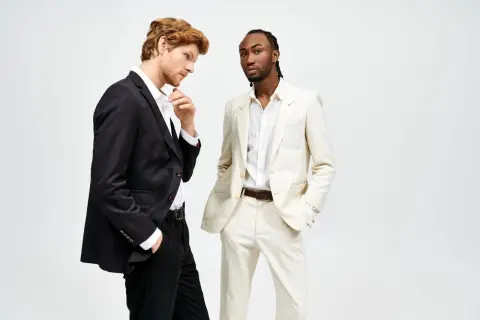 Due uomini in abiti eleganti. Uno con giacca nera e pantaloni neri, l'altro con giacca e pantaloni color crema. Il **slim fit significato** è evidente nella vestibilità moderna.