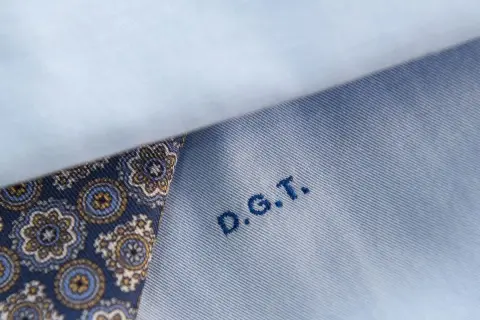 Dettaglio di una camicia azzurra con un inserto di tessuto fantasia blu e oro. Le iniziali D.G.T. sono ricamate in blu.