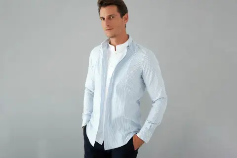 Uomo con outfit camicia a righe azzurre e bianche, indossata aperta su una t-shirt bianca. Mani nelle tasche, look casual.