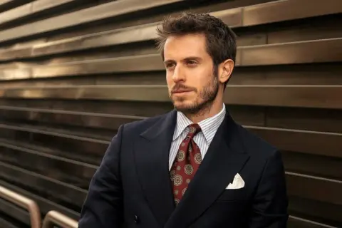 Uomo con abito blu, camicia a righe e cravatta verde con fantasia. Sguardo pensieroso.