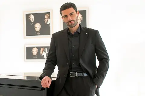 Elegante uomo con camicia invernale uomo nera sotto giacca coordinata, appoggiato a un pianoforte.