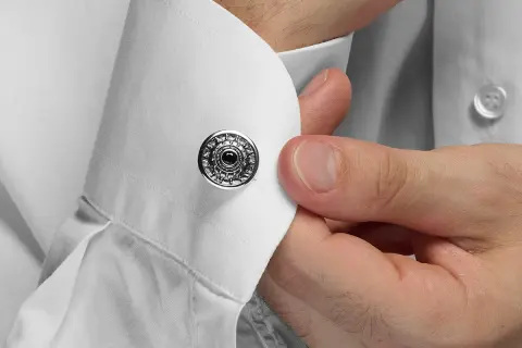Dettaglio su come si mettono i gemelli: una mano inserisce un gemello rotondo con decorazioni argentate e una pietra nera in un polsino di camicia bianca.