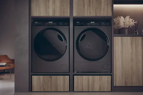 Due asciugatrici nere affiancate, con un cassetto in legno sotto ciascuna. Le lenzuola in asciugatrice sono pronte per essere piegate.