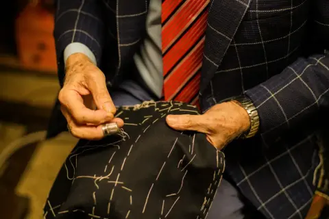 Sarto esperto cuce a mano un tessuto scuro, mostrando i diversi tipi di polsini camicia e la cura artigianale.