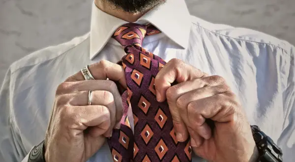 Uomo che sistema il nodo cravatta Eldredge, con camicia bianca e cravatta a rombi arancioni e viola.