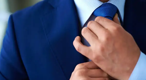 Uomo in abito blu che sistema la cravatta Windsor. Dettaglio elegante e raffinato.
