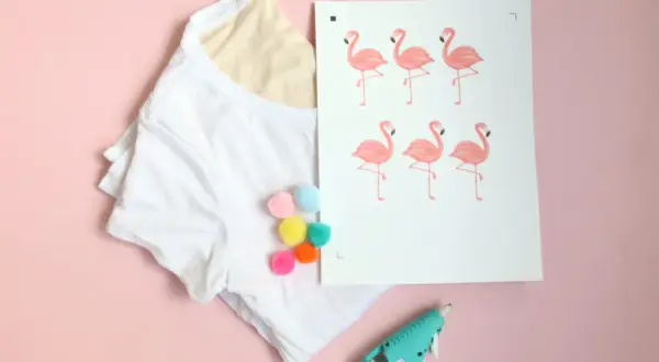 White t-shirt with colorful pom-poms, a print of pink flamingos, and a hot glue gun on a pink background.