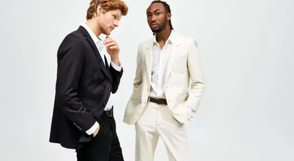 Due uomini in abiti eleganti. Uno con giacca nera e pantaloni neri, l'altro con giacca e pantaloni color crema. Il **regular fit significato** è un taglio classico e confortevole.