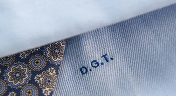 Dettaglio di una camicia azzurra con un inserto di tessuto fantasia blu e oro. Le iniziali D.G.T. sono ricamate in blu.