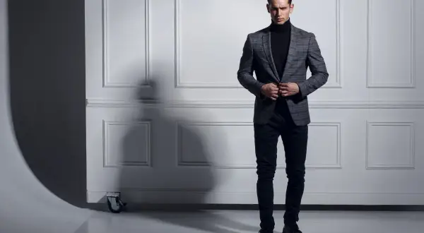 Uomo con maglione nero a collo alto e giacca a quadri grigia, pantaloni scuri e scarpe eleganti.