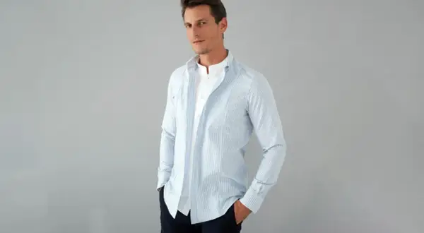 Uomo con outfit camicia a righe azzurre e bianche, indossata aperta su una t-shirt bianca. Mani nelle tasche, look casual.