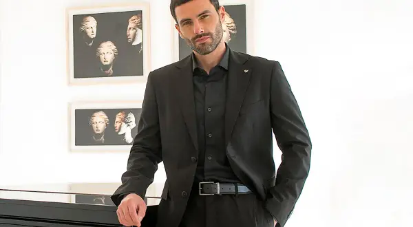 Elegante uomo con camicia invernale uomo nera sotto giacca coordinata, appoggiato a un pianoforte.