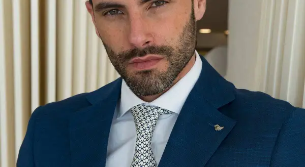 Uomo con vestito blu, camicia bianca e cravatta a fantasia. Sguardo intenso, barba curata.