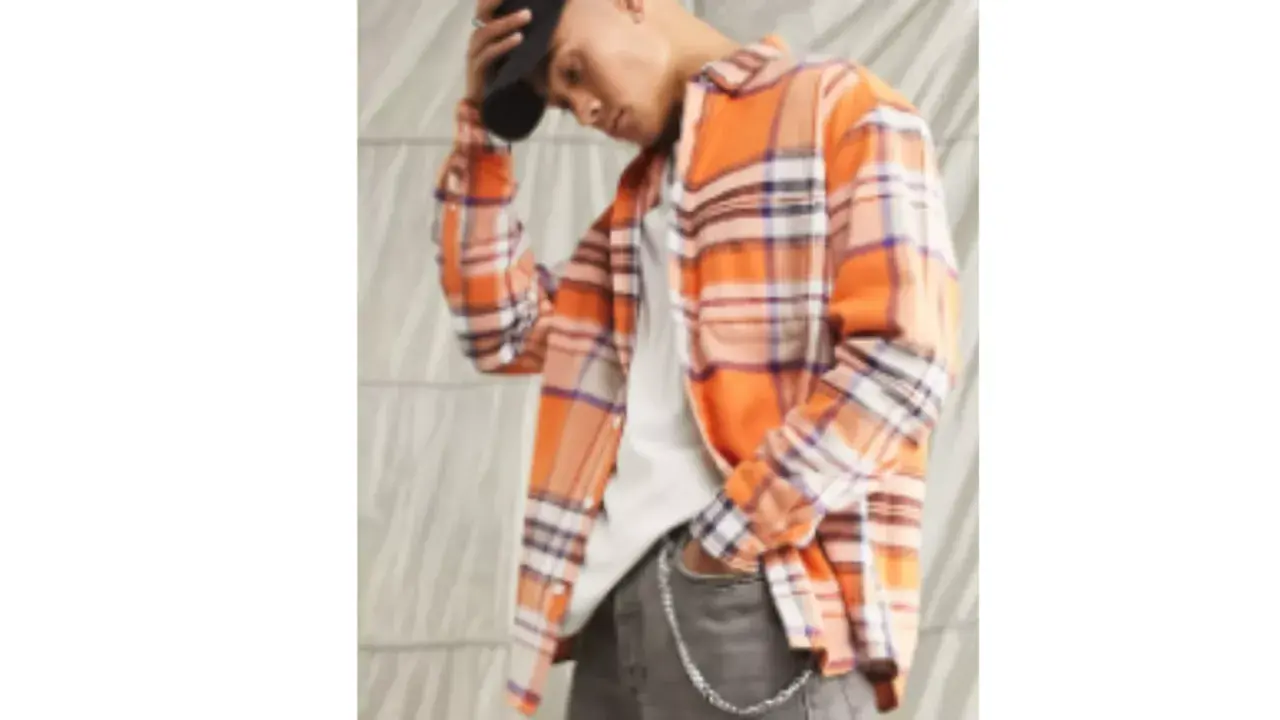 Uomo con camicia in flanella a quadri arancioni, jeans grigi e cappellino nero.