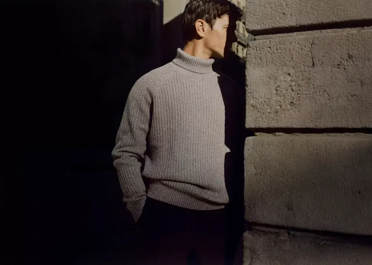 Uomo con un elegante outfit con dolcevita grigio, in piedi vicino a un muro di pietra.