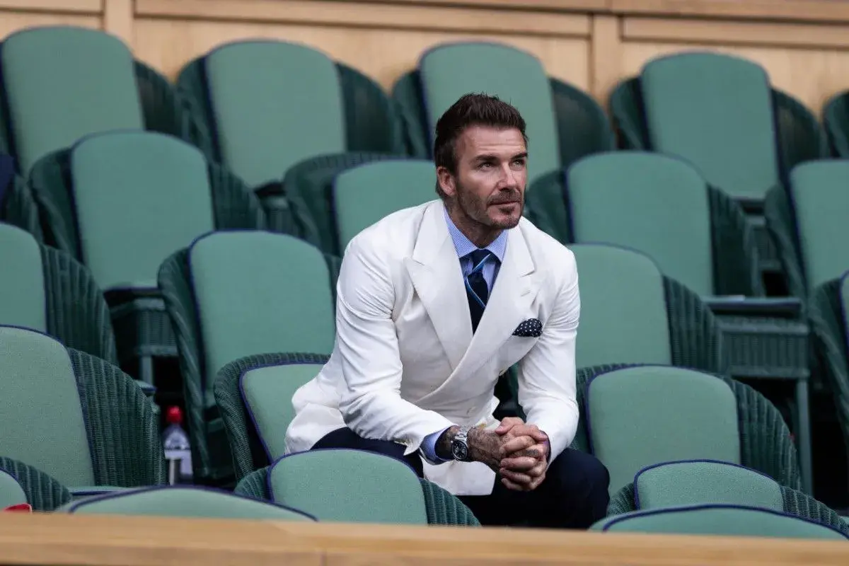 David Beckham in una giacca bianca uomo abbinamento impeccabile, con camicia azzurra e cravatta blu, siede tra le tribune verdi di Wimbledon.