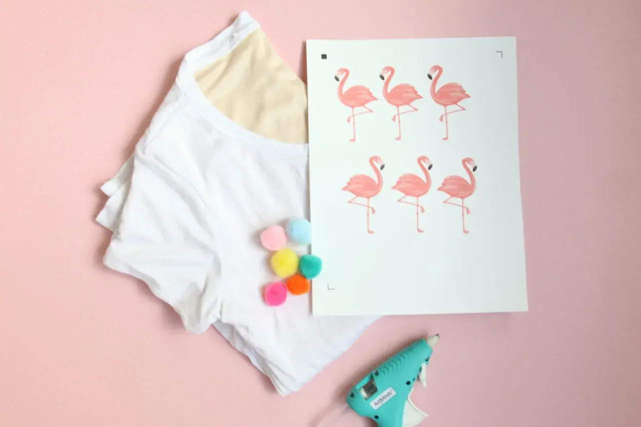 White t-shirt with colorful pom-poms, a print of pink flamingos, and a hot glue gun on a pink background.