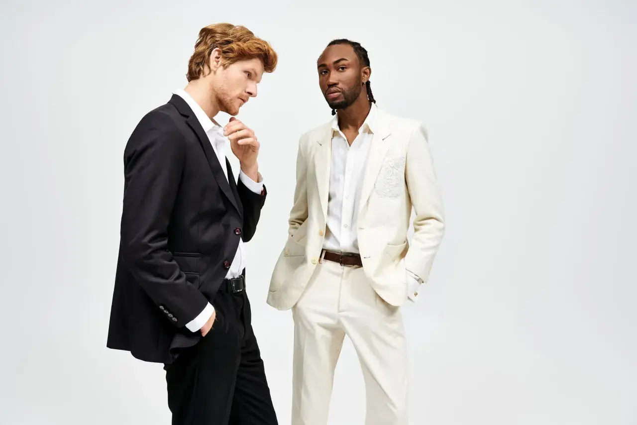 Due uomini in abiti eleganti. Uno con giacca nera e pantaloni neri, l'altro con giacca e pantaloni color crema. Il **regular fit significato** è un taglio classico e confortevole.