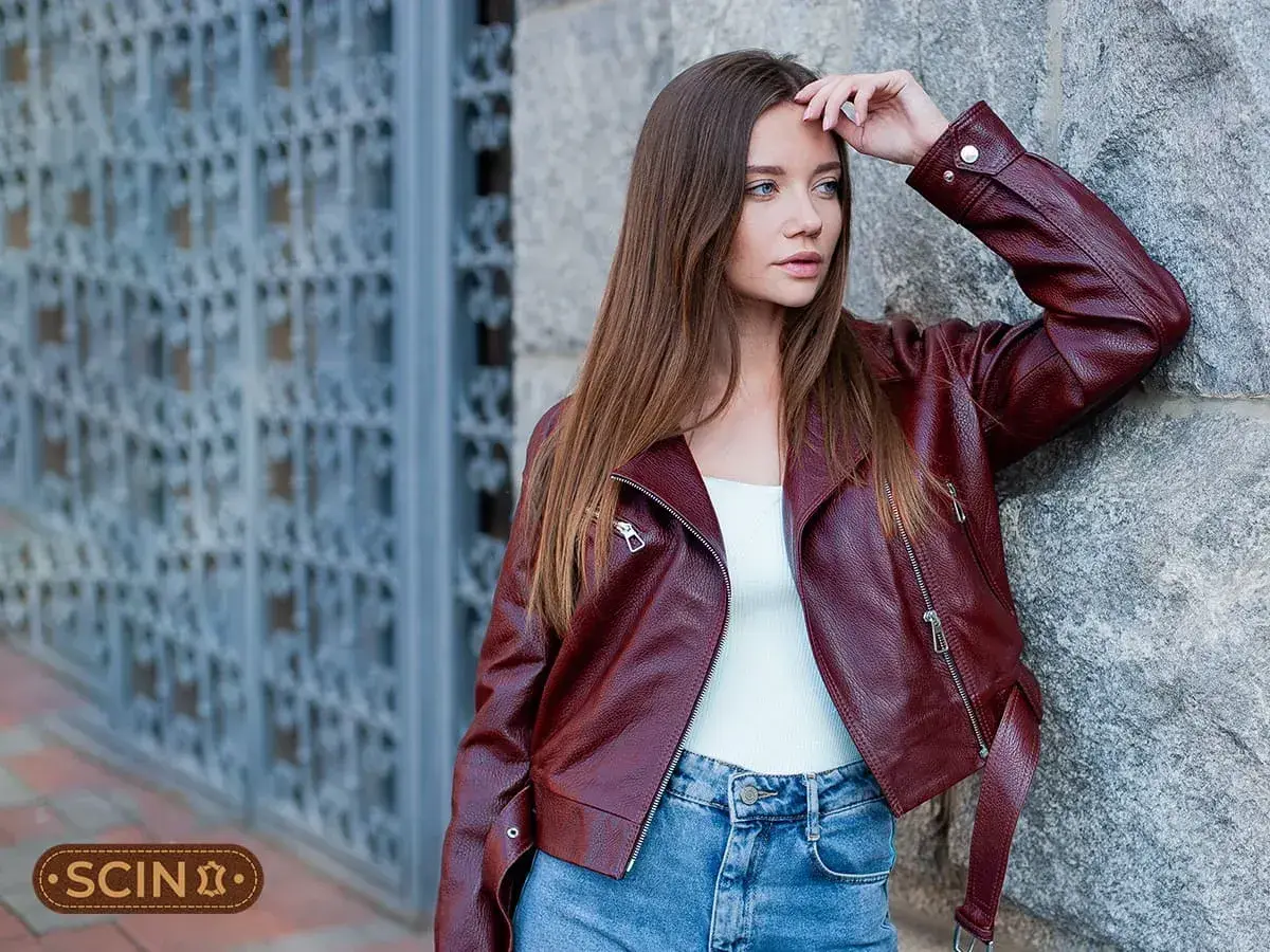 Woman in a burgundy leather jacket and blue jeans leans against a stone wall.