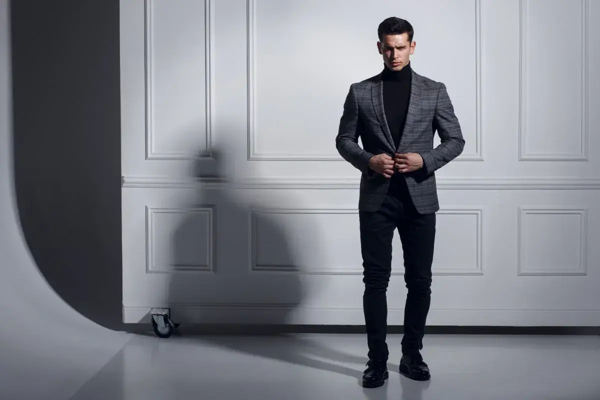 Uomo con maglione nero a collo alto e giacca a quadri grigia, pantaloni scuri e scarpe eleganti.