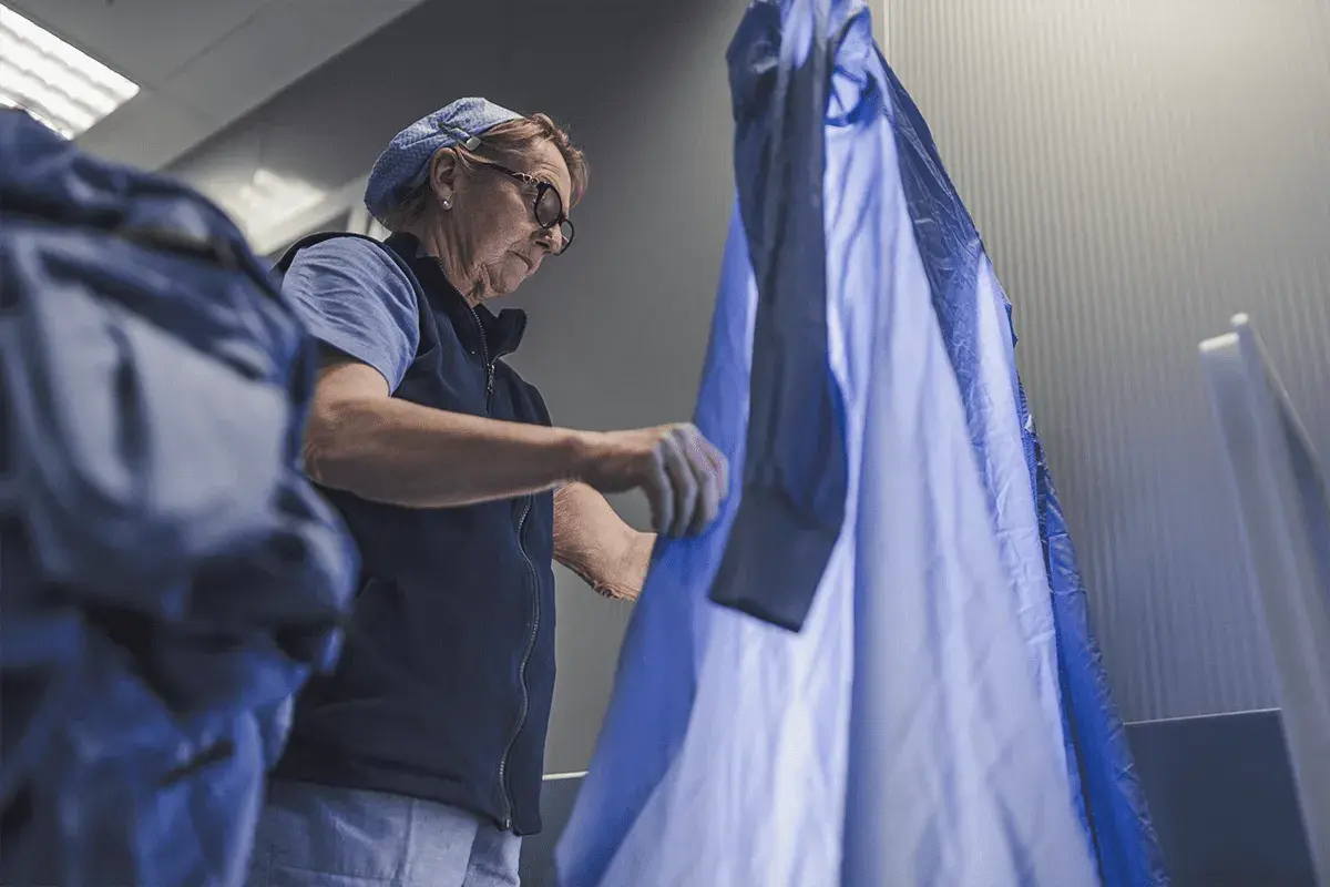 Donna con occhiali e cappellino blu sistema un camice azzurro, forse in una lavanderia.