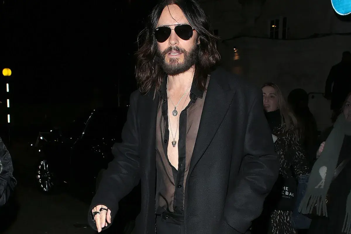 Jared Leto con un audace outfit camicia nera trasparente sotto un cappotto scuro, collane d'argento e occhiali da sole.