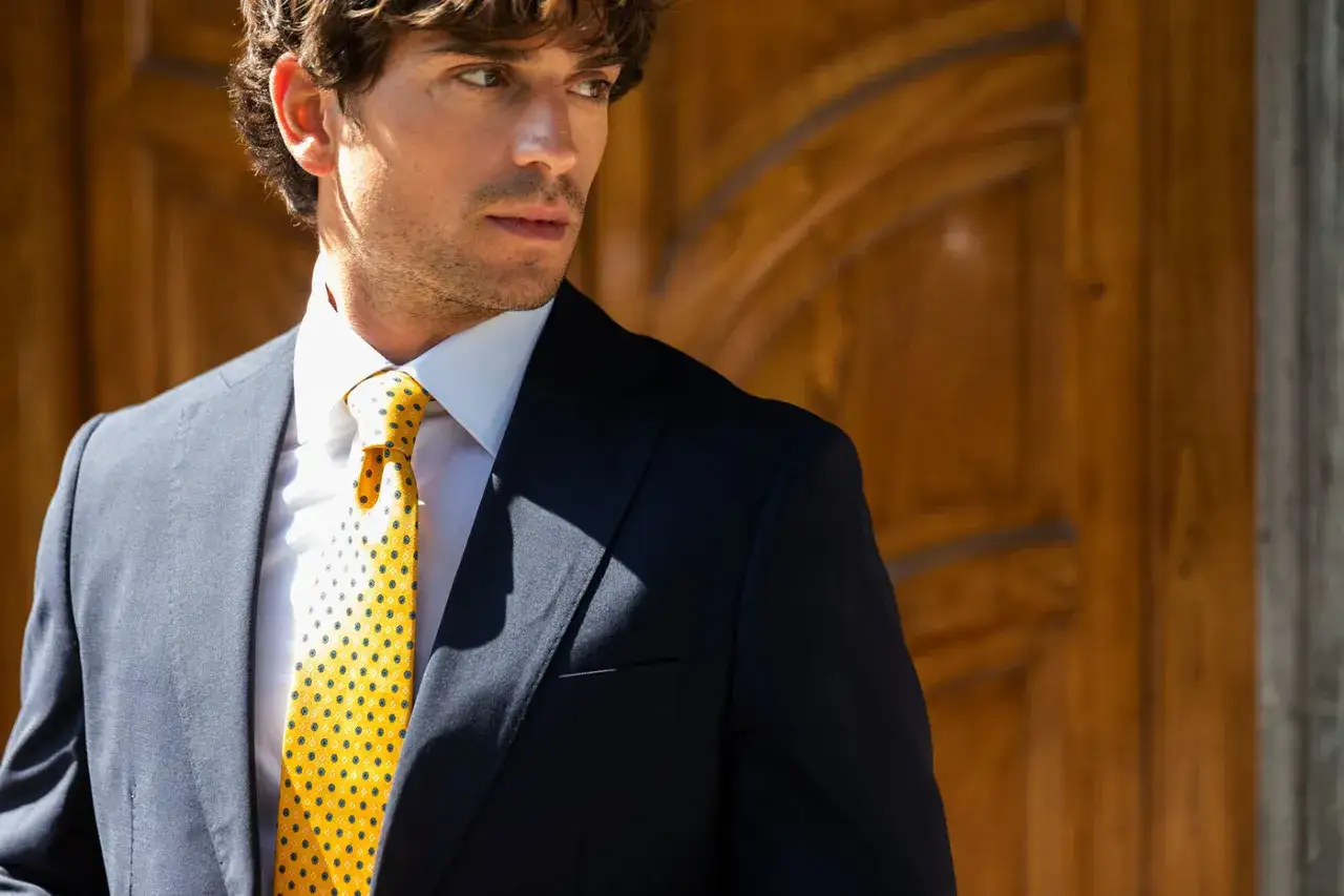 Uomo con giacca blu e camicia bianca, indossa una cravatta gialla a pois blu, un esempio di eleganti tipi di cravatta.