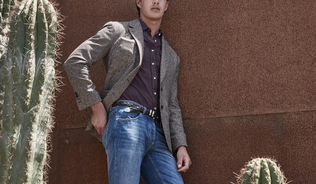 Uomo con un elegante outfit blazer e jeans, appoggiato a un muro arrugginito, con cactus sullo sfondo.