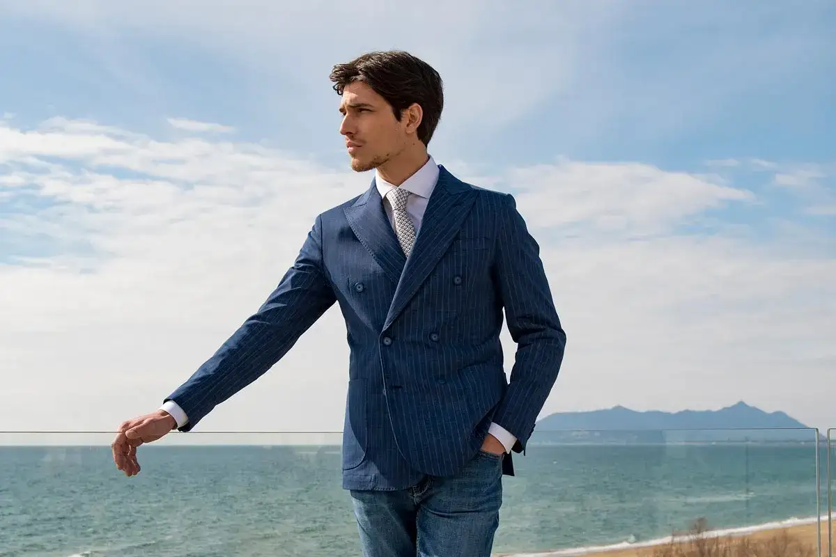 Uomo con giacca doppiopetto blu a righe sottili, camicia bianca e cravatta, con jeans. Il tessuto della giacca ha un leggero effetto color lino, perfetto per una giornata al mare.