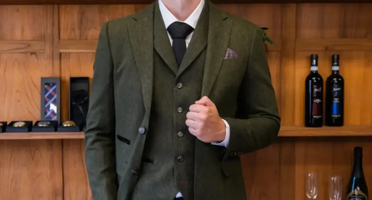 Uomo con abito verde, gilet e cravatta nera. Sullo sfondo, bottiglie di vino e accessori.